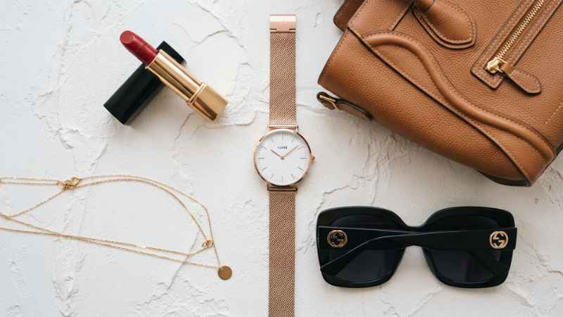 A collection of fashion accessories including a gold watch, sunglasses, and red lipstick arranged on a white surface.