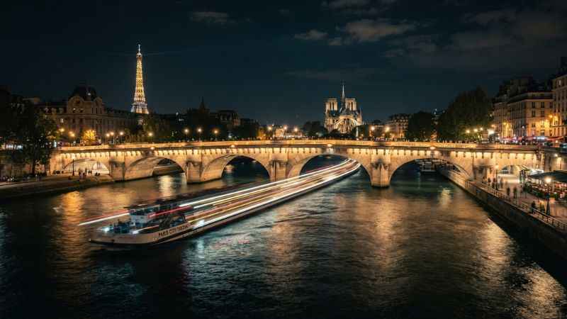 Illuminated bridge over the Seine River with glowing riverboat light trails in Paris during an evening stroll.